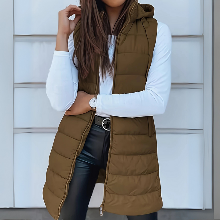 Olive green sleeveless puffer vest with hood worn over white top and black pants, outdoor setting.