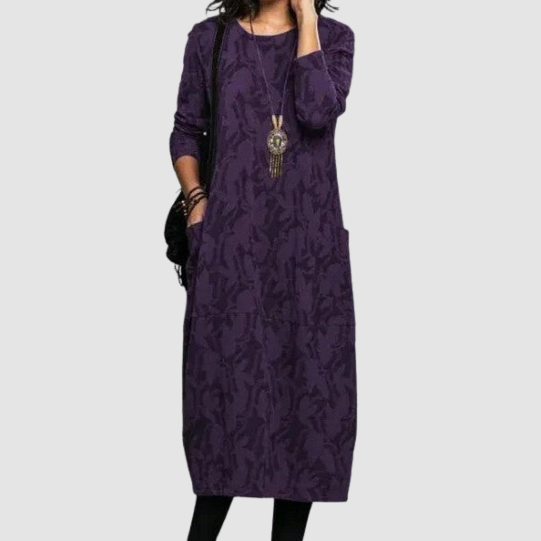 Woman modeling a sophisticated purple dress with leaf motifs, accessorized with a distinctive necklace, on a plain background.