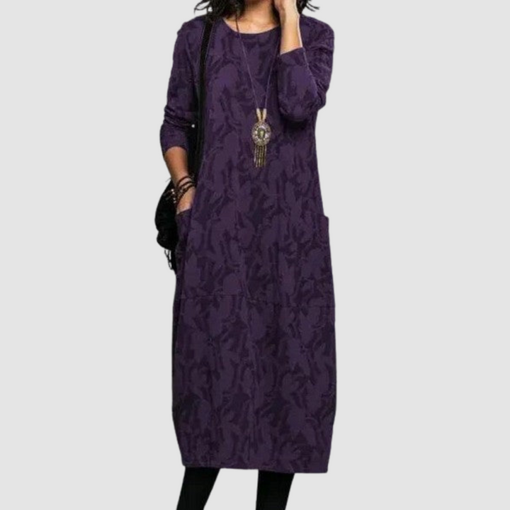Woman modeling a sophisticated purple dress with leaf motifs, accessorized with a distinctive necklace, on a plain background.