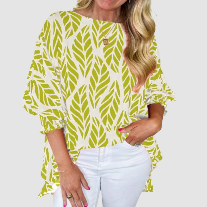 Woman wearing a white and green loose blouse with leaf pattern and white pants in a studio setting.