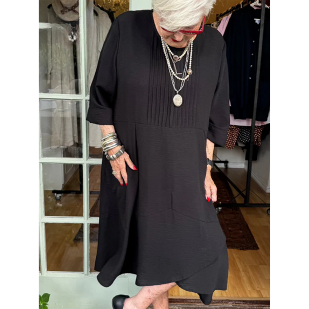 Woman wearing a black pleated dress with irregular hem, accessorized with layered necklaces in a boutique setting.
