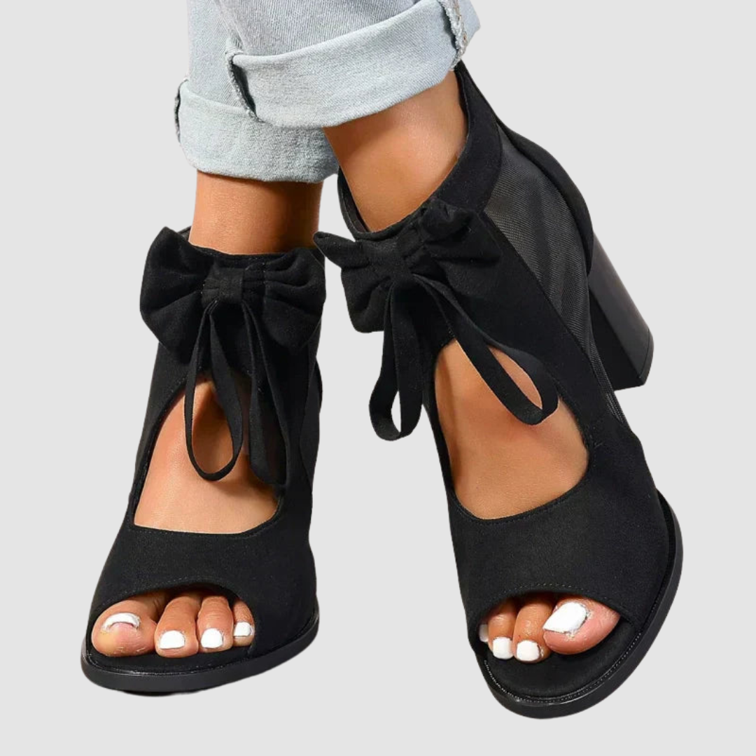 Black heeled sandals with bow detail, worn with cuffed jeans on a neutral background.