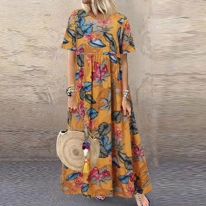 Model in an orange floral dress featuring vibrant leaves, holding a woven bag, standing by a textured wall.