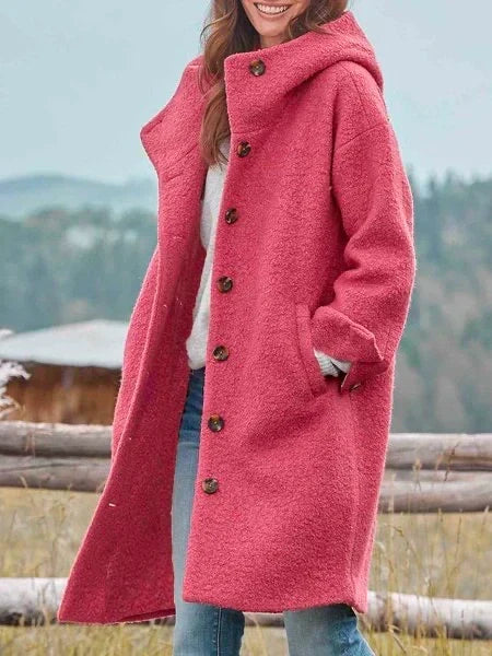 Woman in long pink coat with buttons, standing outdoors near a wooden fence, mountains in background.