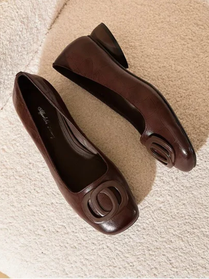 Pair of brown leather flats with buckle detailing, displayed on a textured surface.