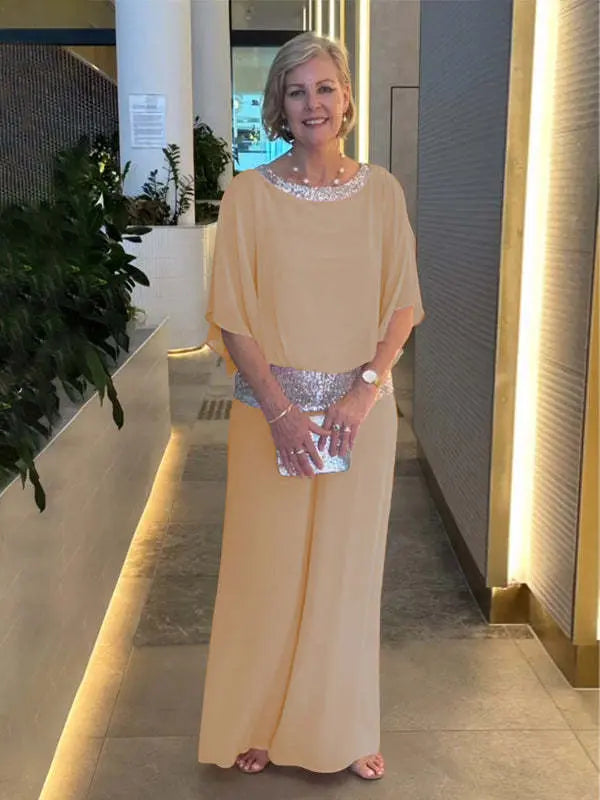 Woman in a light orange elegant set with shimmering belt and neckline, holding a silver clutch. Indoor setting with plants.