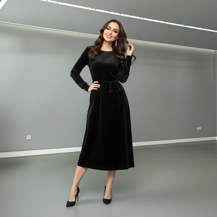 Woman in black suede midi dress with flowing sleeves and belt, posing in sleek, minimalist grey room.