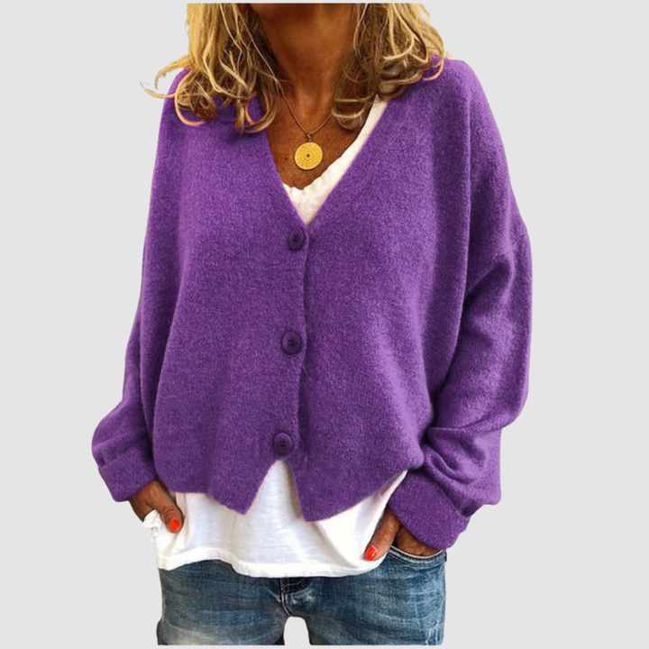 Woman wearing a purple knit cardigan over a white shirt and blue jeans, set against a plain gray background.