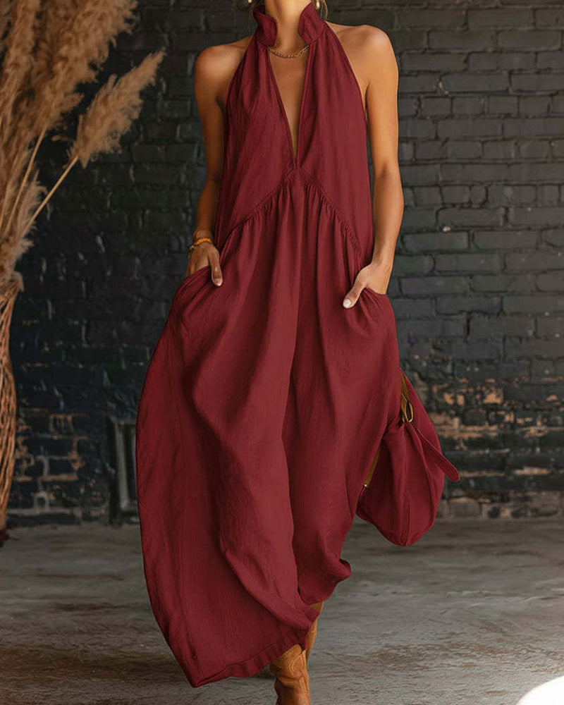 Woman in a deep red maxi dress with a halter neckline, pockets, standing by a rustic brick wall with natural decor.