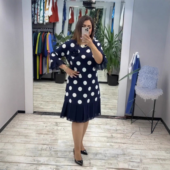 Woman in fitting room mirrors, wearing a navy polka dot pleated dress with white accents, black heels, surrounded by plants and hanging clothes.