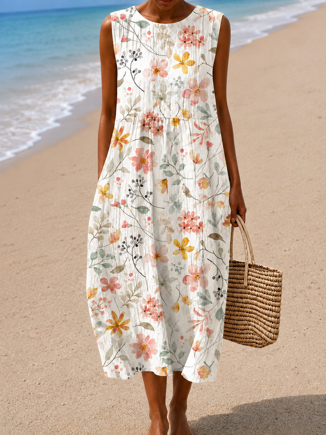 Women’s sleeveless dress with floral patterns, worn on a beach with sand and ocean waves, holding a woven handbag.
