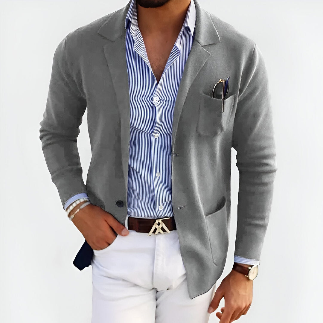 Man wearing a gray lightweight jacket over a striped shirt, paired with white pants, in a studio setting.