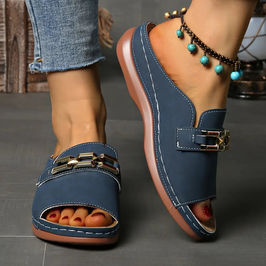 Person wearing blue comfort sandals with gold accents and an anklet, shown in an indoor setting on a grey floor.