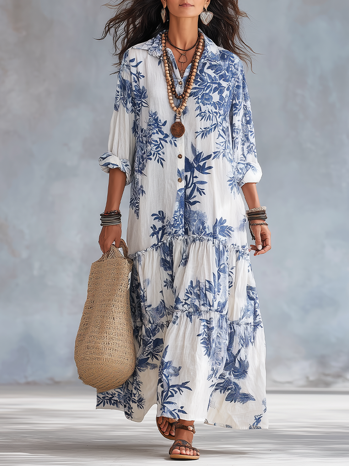 Flowing white maxi dress with blue floral print, worn with layered necklaces and bracelets, holding woven bag in soft studio light.