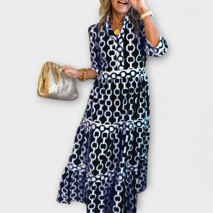 Woman in dark chain print midi dress holding a gold bag against a plain background.