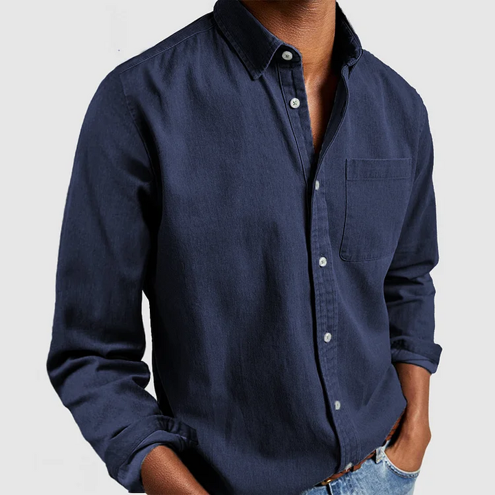 Navy blue button-up shirt with front pocket and rolled sleeves, styled with hands in pockets; gray background.