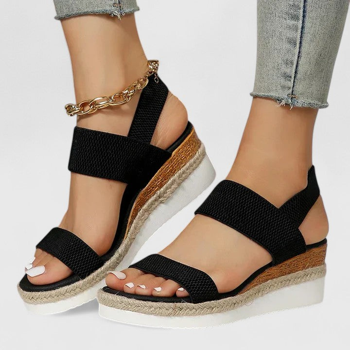 Black orthopedic sandals with thick straps and platform soles, worn with light jeans and a gold anklet in a studio setting.