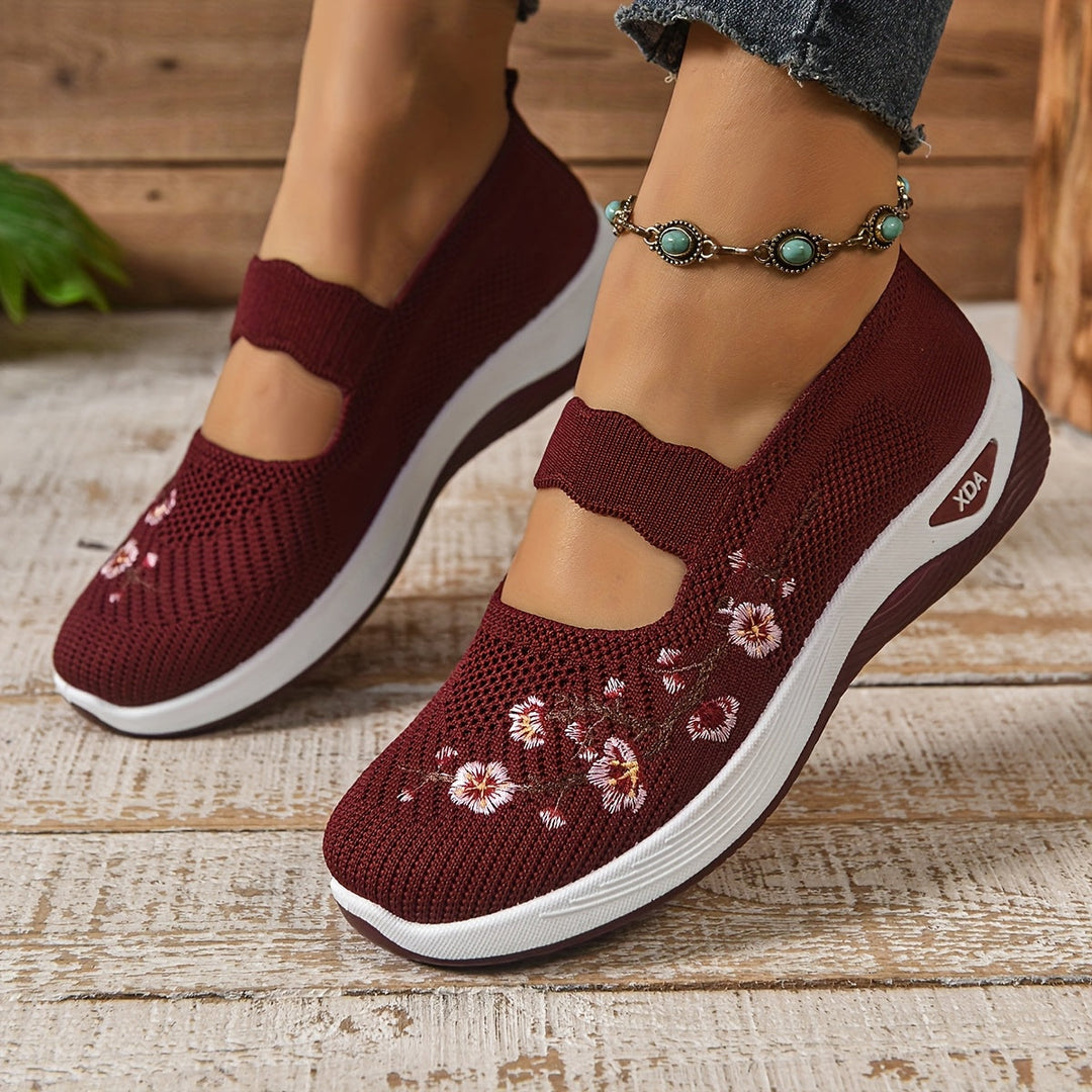 Burgundy orthopedic shoes with floral embroidery, worn with an anklet, on a wooden floor.