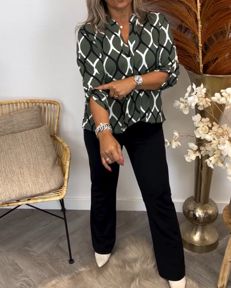 Person adjusting sleeve of a green and white graphic print blouse paired with black pants, in a stylish room with plants and cushions.