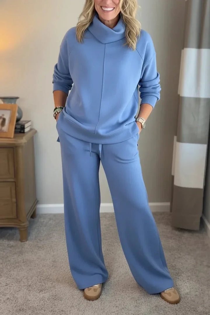 Woman wearing a light blue cozy set with long sleeves and wide-leg pants, standing in a carpeted room beside a wooden table.