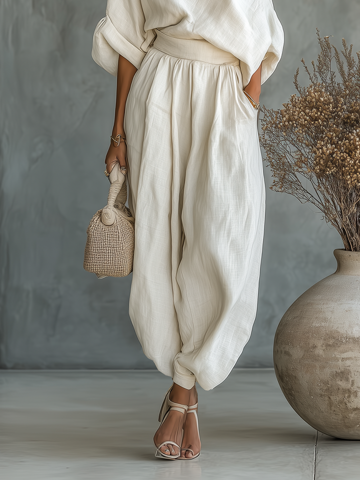 Lower body view of a woman in loose white trousers with heels, holding a beige woven bag indoors.
