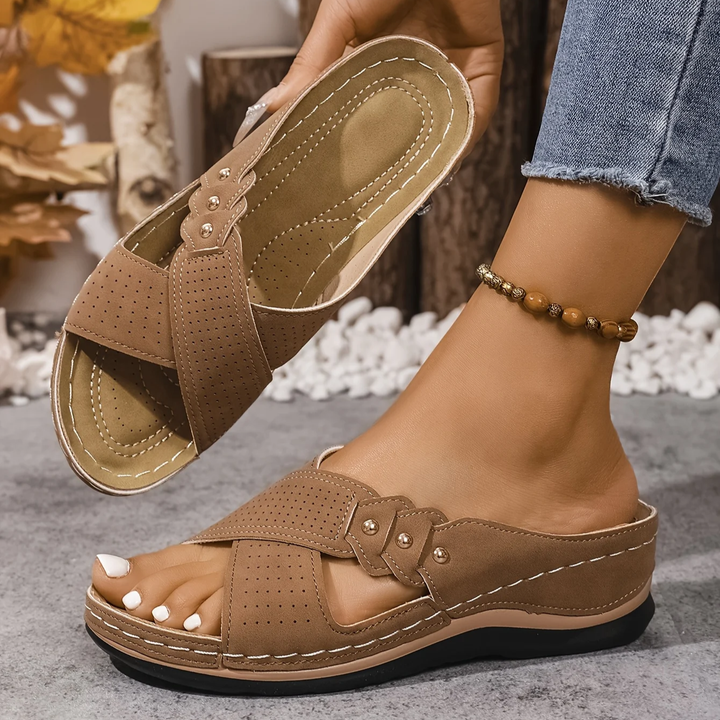 Brown orthopedic wedge sandals held in hand, worn indoors with a nature-inspired backdrop.