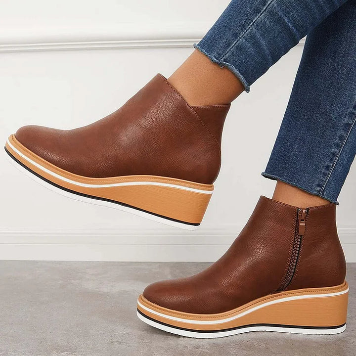 Side view of brown wedge boots with zipper, worn with jeans, against a white wall and gray floor.