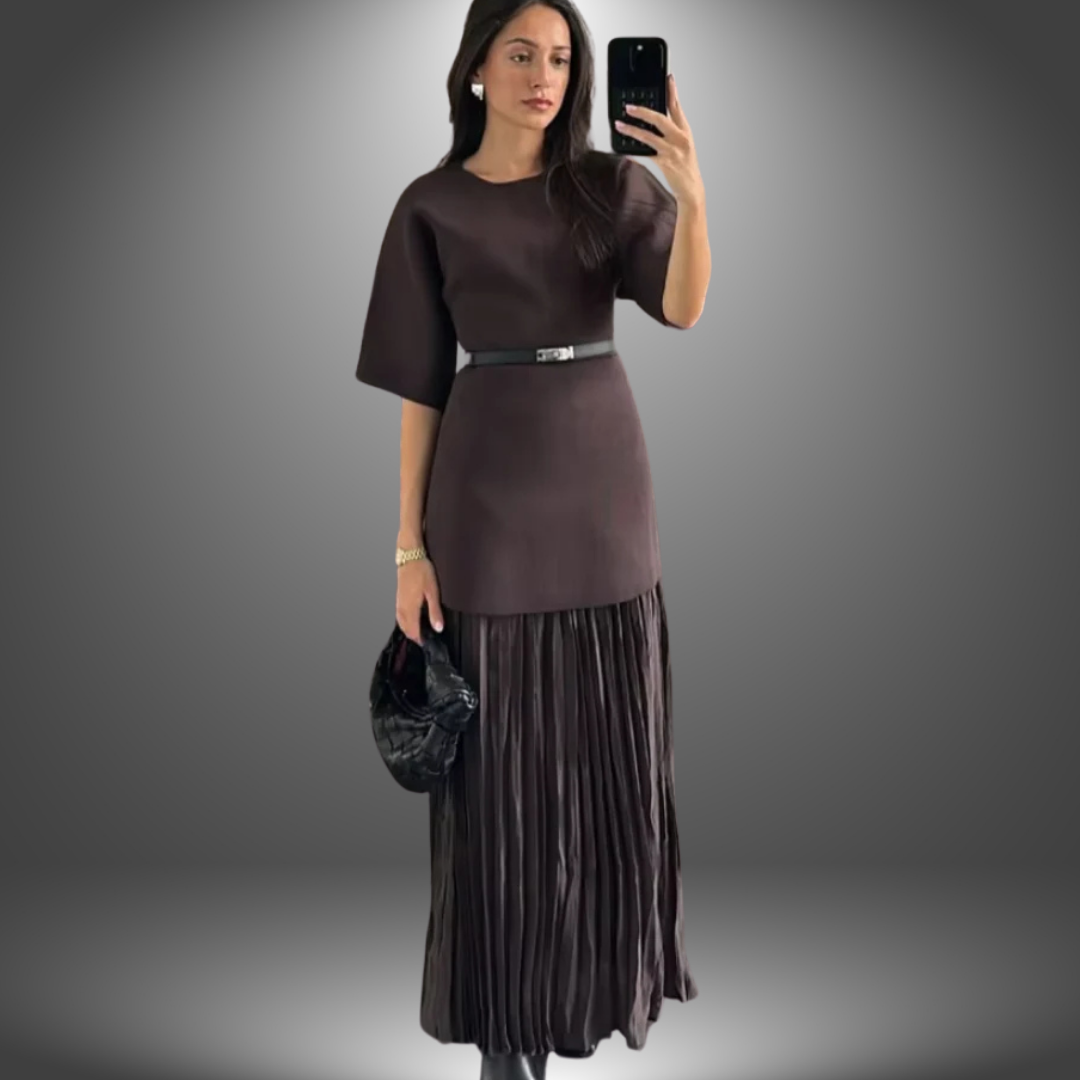 Woman in a dark brown handcrafted dress with short sleeves, pleated skirt, and black belt, posing in a gradient studio background.