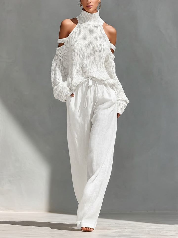Woman dressed in a white turtleneck sweater with cold shoulder cutouts and flowing white pants in a sunlit indoor setting.