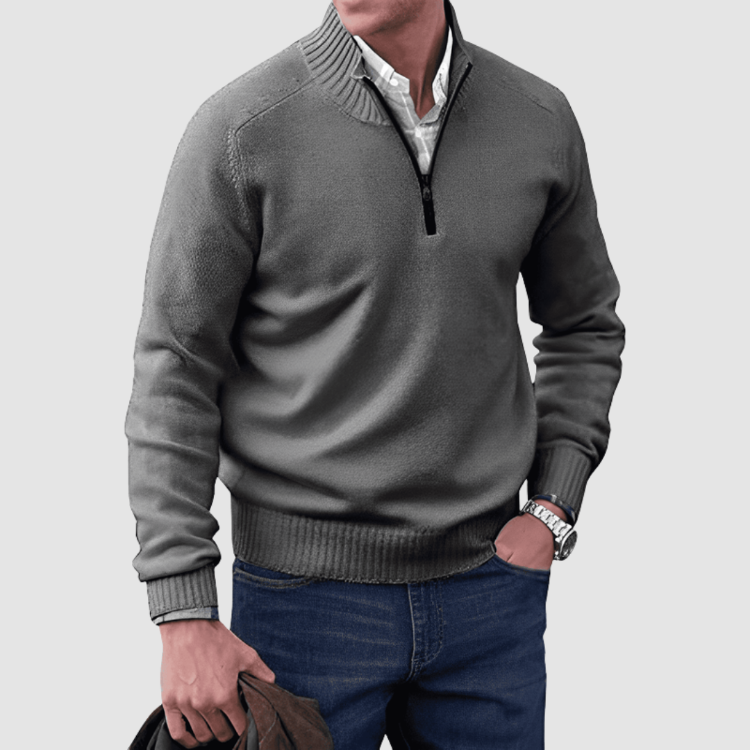 Model in gray zip pullover with ribbed collar and cuffs, holding a jacket. Paired with jeans, set against a plain background.
