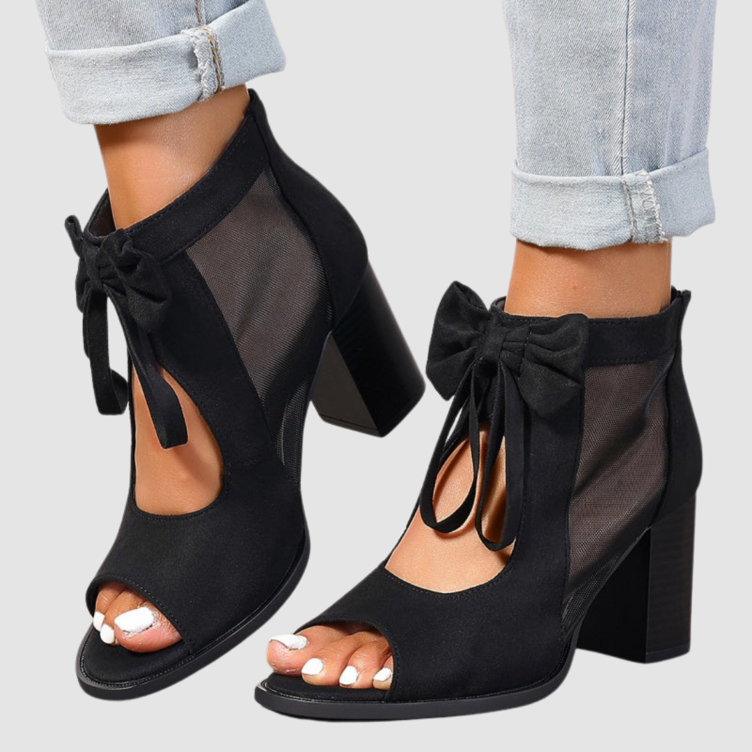 Side view of black heeled sandals with bow and mesh panels, paired with cuffed jeans on a neutral background.