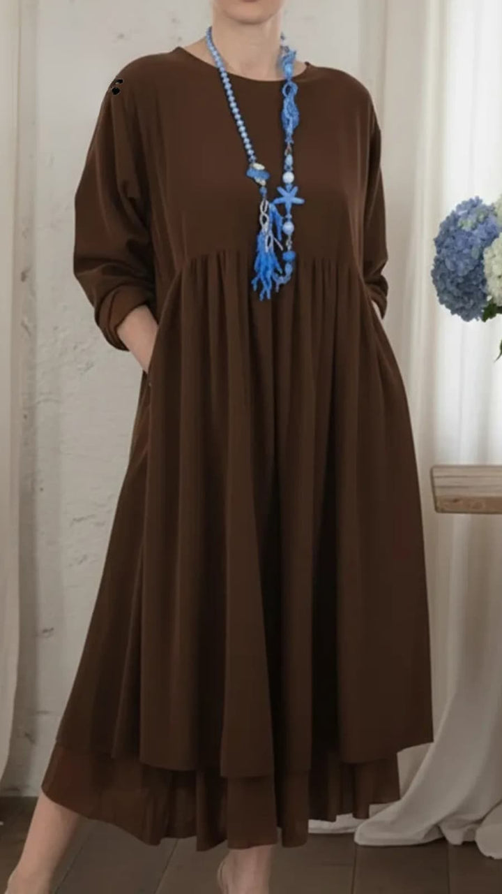 Brown maxi dress with long sleeves, featuring a blue beaded necklace, in an indoor setting with a rustic backdrop and flowers.
