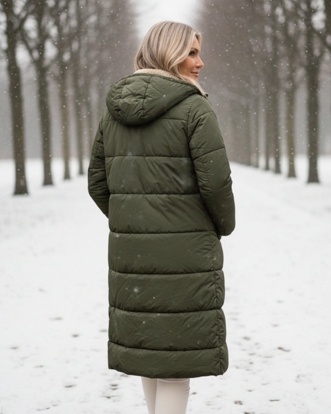 Back view of woman in green coat, walking along a snowy tree-lined path.