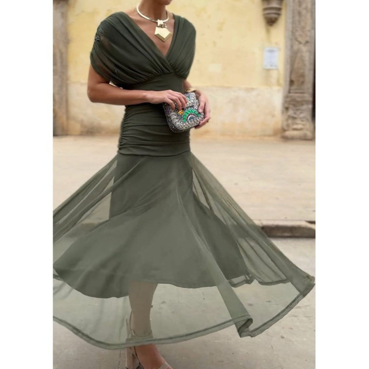 Woman in a dark green mesh dress, holding a decorative clutch, standing outdoors with a rustic wall in the background.