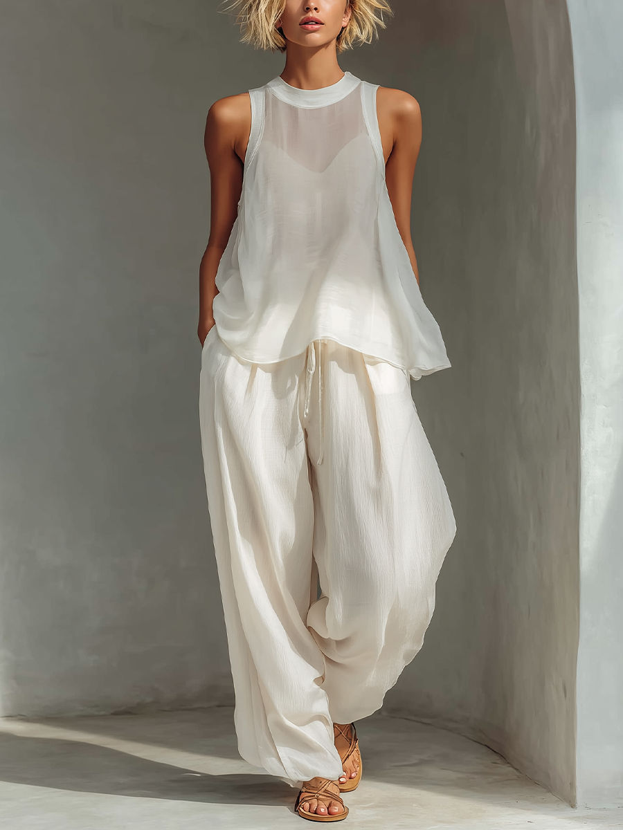 Model in stylish white outfit with sheer top and wide-legged pants in light-filled, minimalist space.