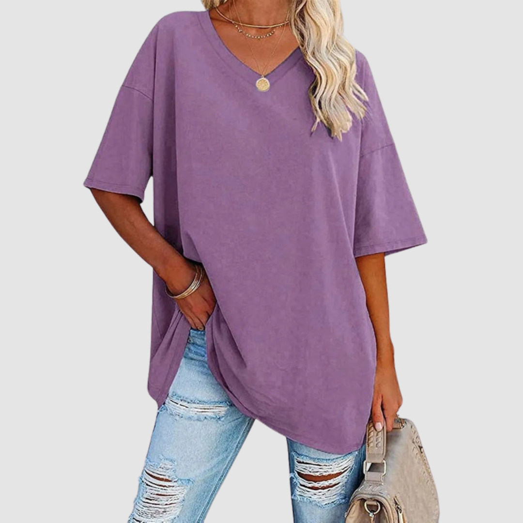 Woman in a purple V-neck T-shirt with distressed jeans, carrying a beige bag, studio background.