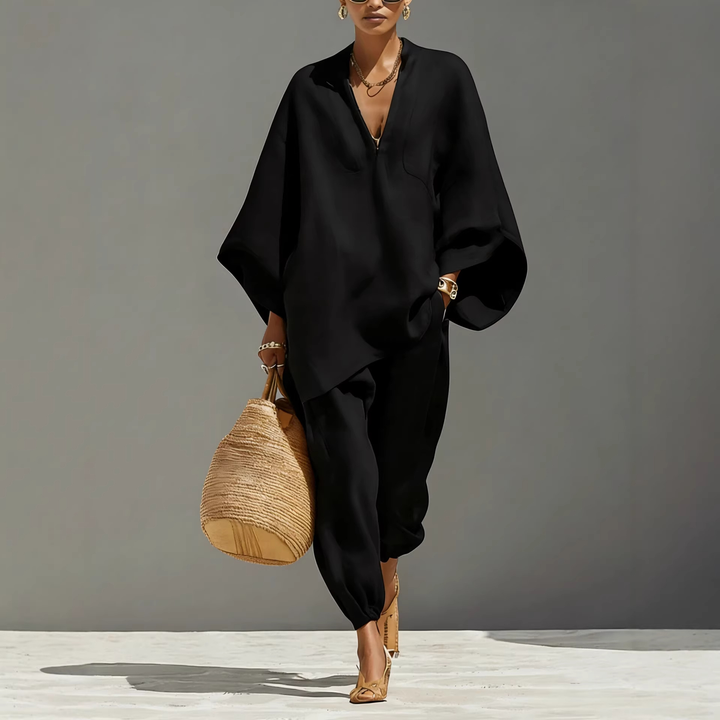 Model in black batwing sleeve V-neck lounge set carrying woven bag, studio background.