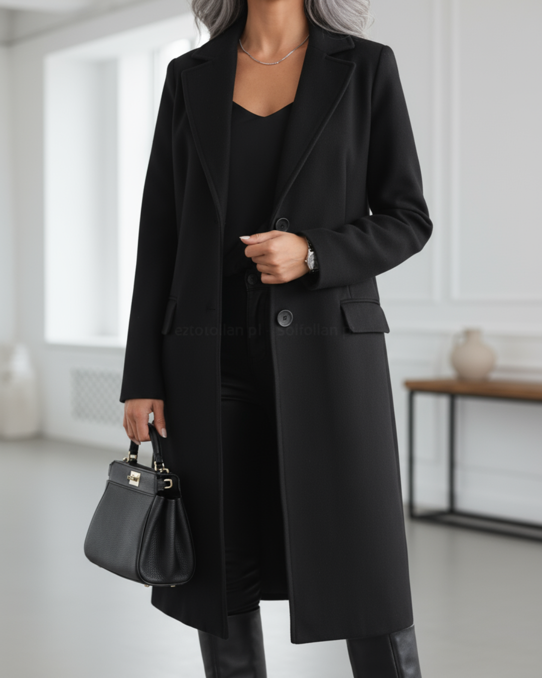 Black long coat with button front, worn over black attire, paired with a handbag in a stylish, modern interior.
