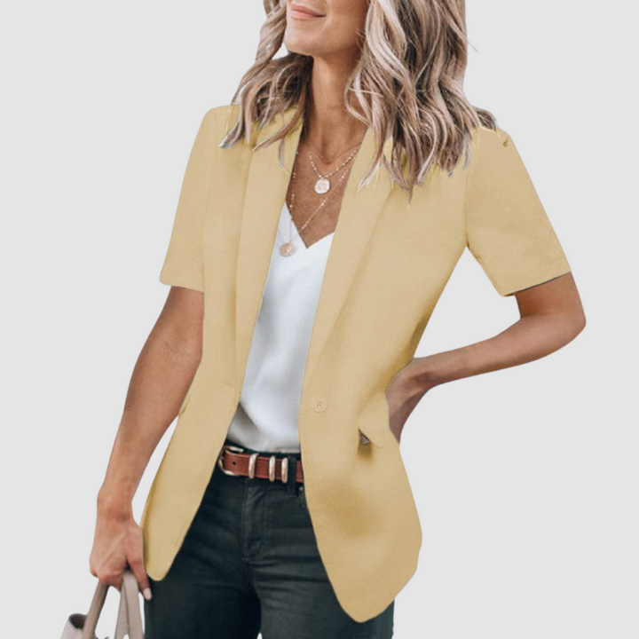 Yellow short-sleeve blazer worn by person with wavy hair, white top, and jeans; holding a beige handbag against a plain background.