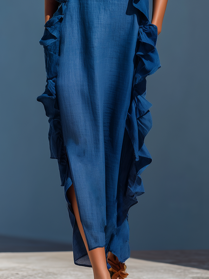 Lower half of blue maxi dress with ruffles and side slit, worn with strappy sandals, against a blue wall background.
