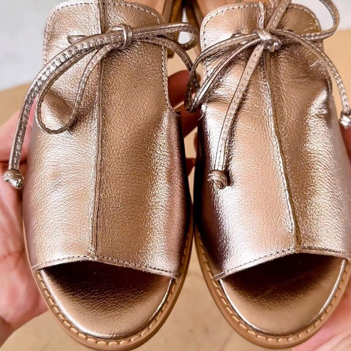 Close-up of metallic leather sandals with bow detail on a wooden surface.