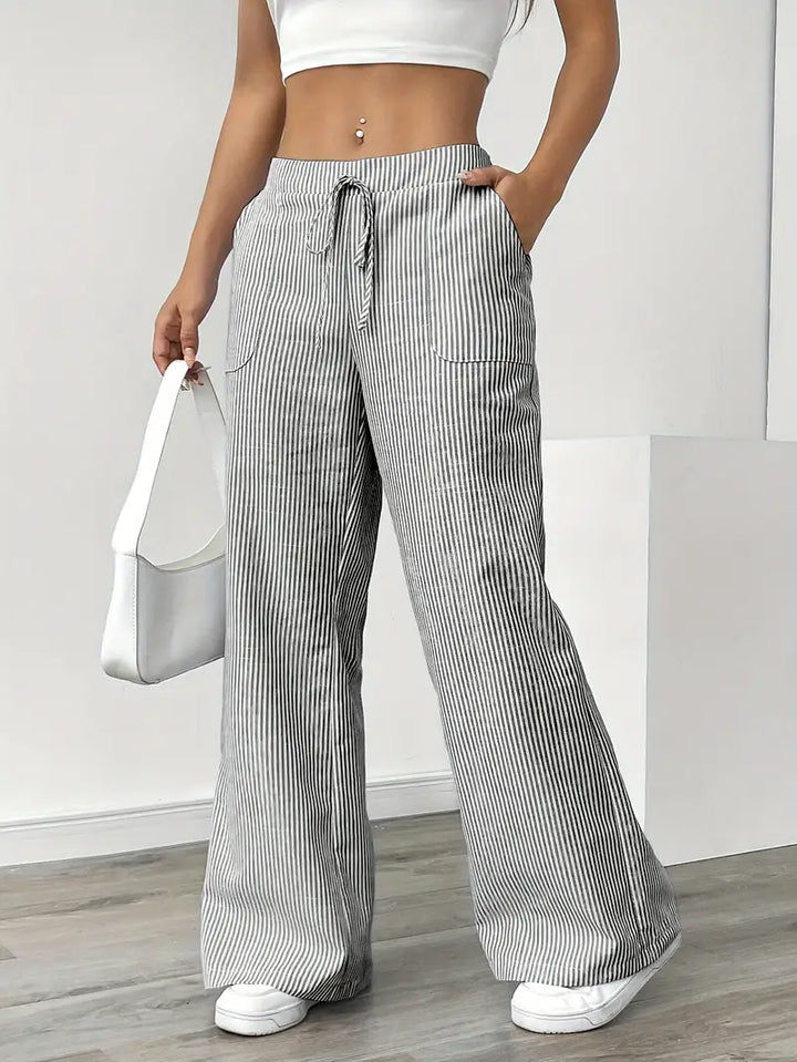 Gray and white striped pants with pockets and drawstring, modeled indoors with white sneakers and white bag.