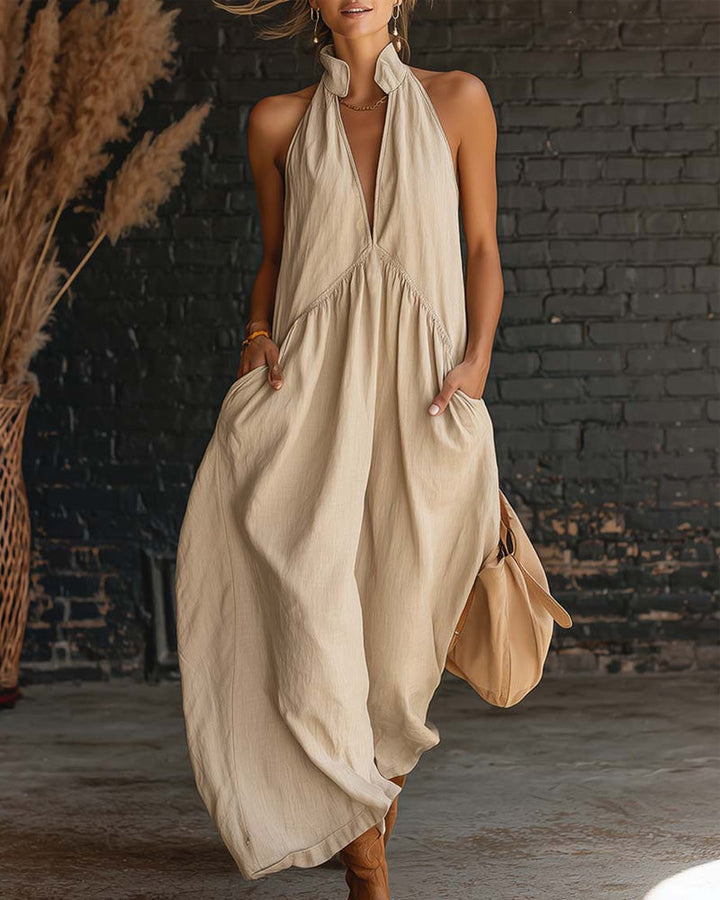 Woman in beige maxi dress with halter neckline and pockets, posed in front of a textured brick wall and dried grass.