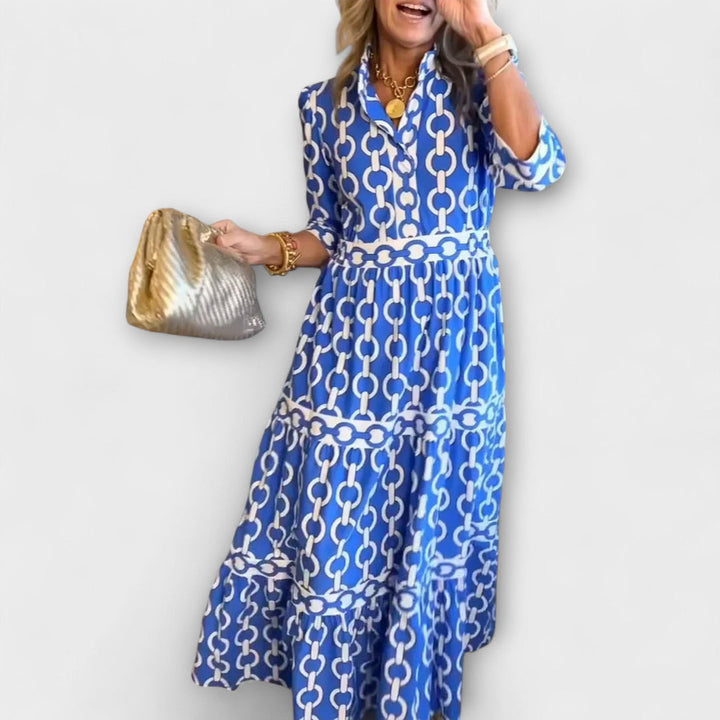 Woman in blue and white chain print midi dress holding a gold bag against a plain background.