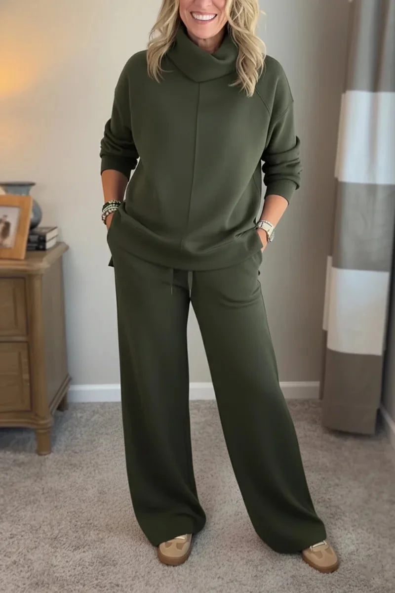 Woman in a green cozy set with long sleeves and wide-leg pants, standing in a carpeted room beside a wooden table.