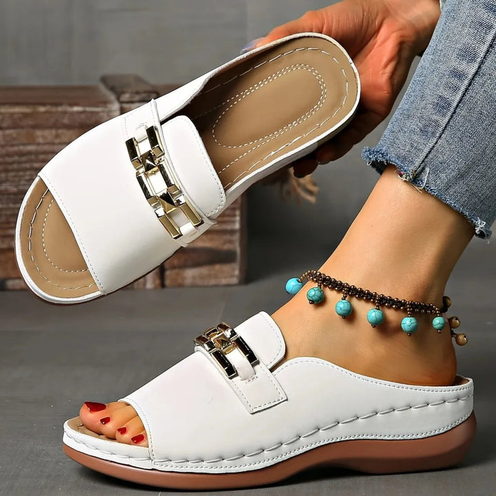 White comfort sandals with gold details, presented on feet with blue anklet, shown in a rustic indoor setting.