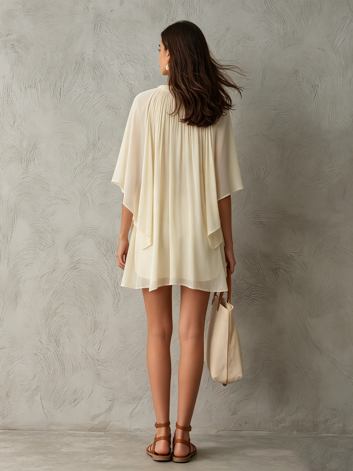 Back view of cream mini dress with flowing cape sleeves, paired with sandals and tote bag, textured wall backdrop.