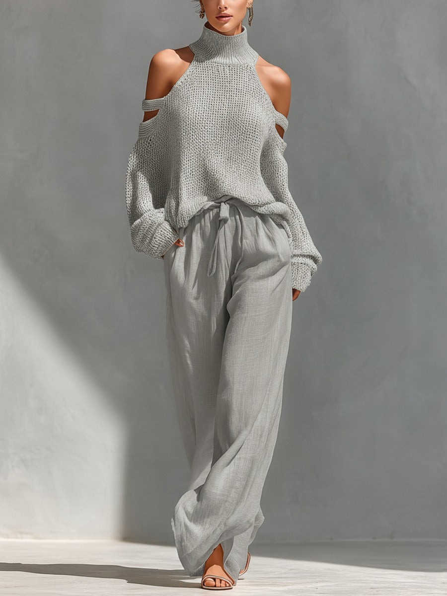 Woman in a light grey turtleneck sweater with cold shoulder cutouts and flowing grey pants in a sunlit indoor setting.