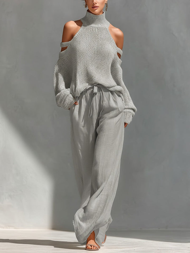Woman in a light grey turtleneck sweater with cold shoulder cutouts and flowing grey pants in a sunlit indoor setting.