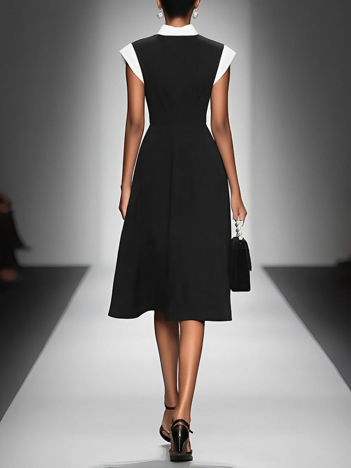 Back view of model in black midi dress with white collar, holding handbag, walking down runway, studio lights.